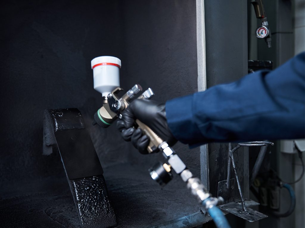 A person in a blue sleeve and black glove uses a spray gun to apply advanced coatings, ensuring industrial durability as they coat a metal object with black paint inside a spray booth. - Alwande Tech