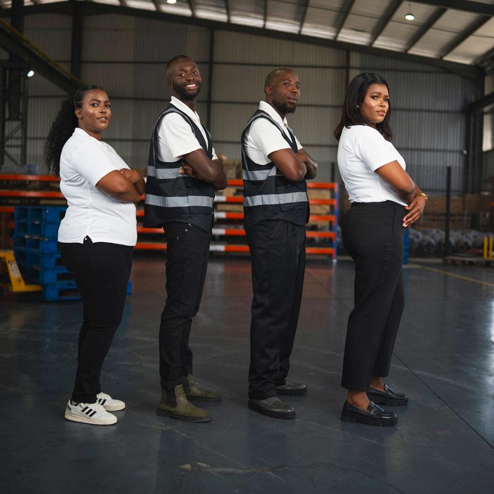 Four warehouse workers stand in a row with arms crossed, wearing uniforms and safety vests, inside an industrial storage facility—showcasing the team spirit at AlwandeTech South Africa. - Alwande Tech