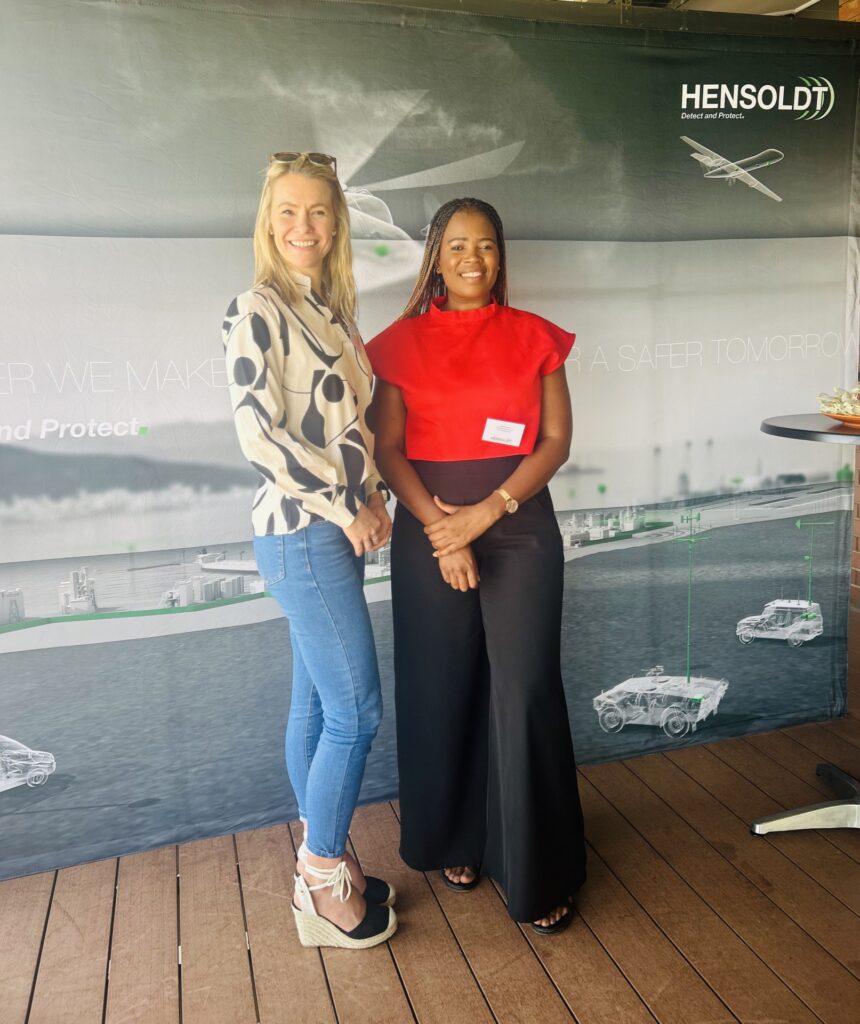 Two women stand and smile in front of a HENSOLDT promotional backdrop at an indoor event. - Alwande Tech