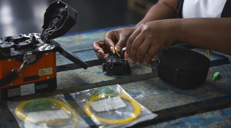 A person works on a fibre optic cable with tools on a blue wooden table; two yellow cables in plastic bags are visible in the foreground. - Alwande Tech
