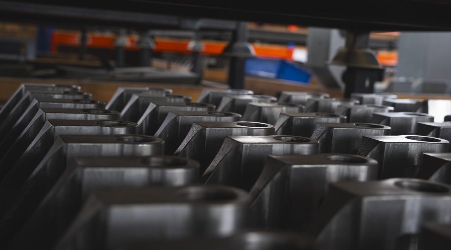 Rows of metal machine parts are neatly arranged in an industrial setting, with blurred machinery and shelving visible in the background. - Alwande Tech